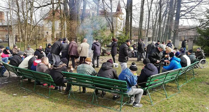 Mieszkańcy Domu Pomocy Społecznej w Wieleniu siedzą w parku na ławkach wokół ogniska, w którym smażą się kiełbaski.
