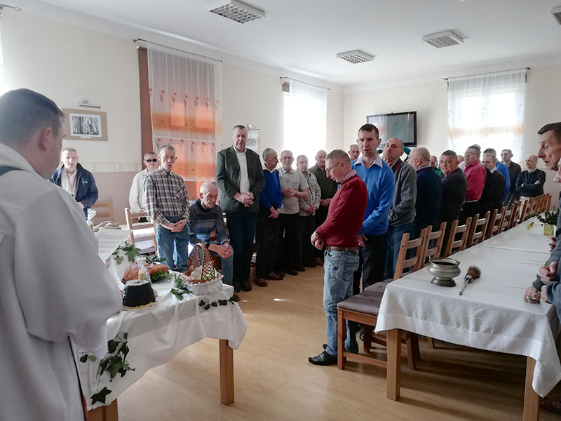 Mieszkańcy Domu Pomocy Społecznej w Wieleniu stoją przy stołach wielkanocnych podczas święcenia pokarmów przez księdza.
